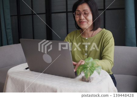 Senior women using laptop at home, sitting alone on sofa and smiling. 139448428