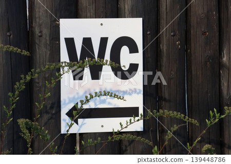 Outdoor WC Direction Sign Against Wooden Fence with Nature Elements for Rustic Decor 139448786