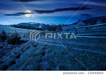 mountainous countryside landscape in spring at night. full moon over wooden fence and grassy meadow on a green hill. distant mountain ridge with snow capped tops. mysterious cloudy sky 139448844