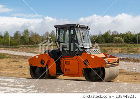 Asphalt paver is at a construction site laying asphalt on a road while surrounded by trees and open land under a clear sky Asphalt paver is at a construction site laying asphalt on a road while surrounded by trees and open land under a clear sky 139449233