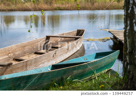 Two boats sit next to a wooden pier at the edge of a body of water with grass and trees nearby. The sun shines on the scene Two boats sit next to a wooden pier at the edge of a body of water with grass and trees nearby. The sun shines on the scene 139449257