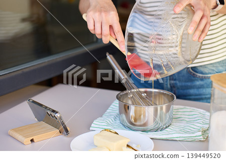 Woman pours batter from glass bowl into saucepan with whisk beside smartphone Woman pours batter from glass bowl into saucepan with whisk beside smartphone 139449520