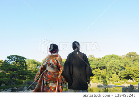 一對身著傳統日式服飾的新郎新娘，背對著鏡頭，仰望著日式庭院中的藍天。背景留有大片天空圖案的文字空間 | 日式婚禮廣告/婚禮橫幅素材 139453245