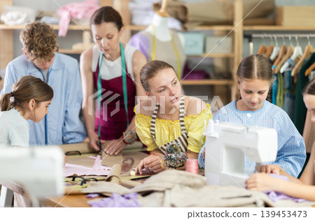 Teen girl watching peer sewing at machine during class Teen girl watching peer sewing at machine during class 139453473