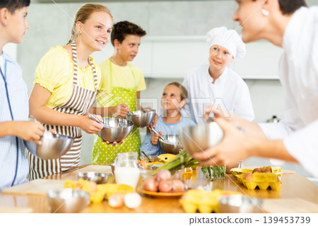 While cooking classes, teen girl stands on other side of table from cook guy and looks at him While cooking classes, teen girl stands on other side of table from cook guy and looks at him 139453739
