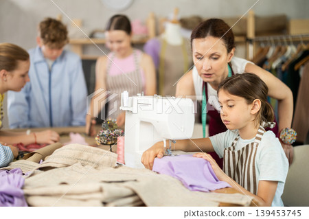 Teenage girl tries to sew on sewing machine. Teacher helps. Children watch and learn Teenage girl tries to sew on sewing machine. Teacher helps. Children watch and learn 139453745