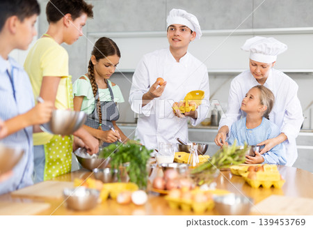 During cooking lesson, male cook tells children about culinary rules, woman assistant helps child During cooking lesson, male cook tells children about culinary rules, woman assistant helps child 139453769