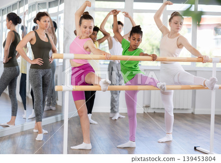 Teenage girls doing leg stretching exercise holding by ballet barre during dance courses Teenage girls doing leg stretching exercise holding by ballet barre during dance courses 139453804