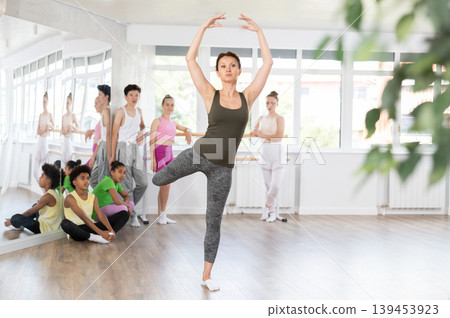 Gracious middle-aged female trainer demonstrating professional ballet poses to teenage boys and girls Gracious middle-aged female trainer demonstrating professional ballet poses to teenage boys and girls 139453923