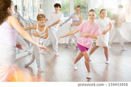 Female teacher performs choreographic exercises and teaches energetic mobile house dance together with teenagers Female teacher performs choreographic exercises and teaches energetic mobile house dance together with teenagers 139453956