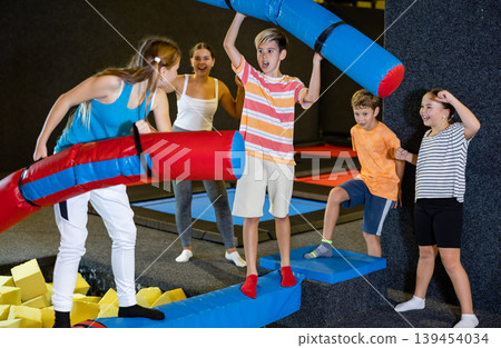 Cheerful school-age boy and teen girl friends having fun on inflatable gladiator fight arena in indoor amusement center Cheerful school-age boy and teen girl friends having fun on inflatable gladiator fight arena in indoor amusement center 139454034