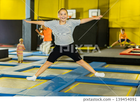 Full length of happy casual girl jumping spreading legs on colorful trampoline at playground park and smiling at camera Full length of happy casual girl jumping spreading legs on colorful trampoline at playground park and smiling at camera 139454091