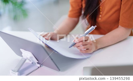 Asian businesswoman writing notes on paper document while reviewing work on digital tablet at modern office desk remote work from home hybrid workplace concept 139455298