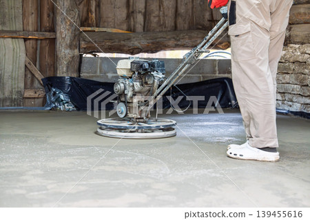 Construction worker operating a power trowel to finish and smooth a newly poured concrete floor Construction worker operating a power trowel to finish and smooth a newly poured concrete floor 139455616
