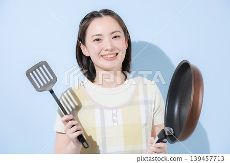 一位家庭主婦身穿圍裙,手持煎鍋,站在淺藍色背景前,面帶燦爛的笑容,直視鏡頭,她可能是家政幫工,也可能是全職家庭主婦,正在做飯。 一位家庭主婦身穿圍裙,手持煎鍋,站在淺藍色背景前,面帶燦爛的笑容,直視鏡頭,她可能是家政幫工,也可能是全職家庭主婦,正在做飯。 139457713