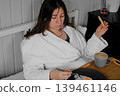 A calm woman enjoys a quiet breakfast. A serene lady sits in bed in a robe, holding a mug and a cracker, in the cozy atmosphere of her home. Morning rituals alone. A cozy interior. Copy space. Banner 139461146