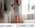 A woman blow-drying her hair, soft morning sunlight shining through the curtains, a concentrated expression on her face, hands running through chestnut curls, a home routine, relaxed self-care. 139461148