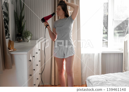 A woman blow-drying her hair, soft morning sunlight shining through the curtains, a concentrated expression on her face, hands running through chestnut curls, a home routine, relaxed self-care. A woman blow-drying her hair, soft morning sunlight shining through the curtains, a concentrated expression on her face, hands running through chestnut curls, a home routine, relaxed self-care. 139461148