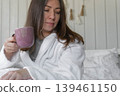A woman lies on a pastel-colored bed, holding a coffee mug. A calm woman in a robe enjoys the serene morning atmosphere at home. Morning routine. Self-care. Starting a new day. Space for text. Banner 139461150