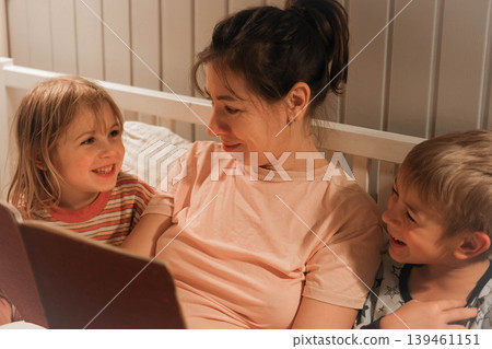 Family bedtime stories. A mother reads a story to her children, wrapped in a blanket. Children listen to soothing stories. A cozy family scene before bed in a softly lit bedroom. Copy space. Banner Family bedtime stories. A mother reads a story to her children, wrapped in a blanket. Children listen to soothing stories. A cozy family scene before bed in a softly lit bedroom. Copy space. Banner 139461151