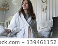Woman in white robe on bed, disheveled hair, soft sunlight through window, clock on nightstand showing morning time, pensive freelancer getting ready for a new day, neutral tones in bedroom. Banner 139461152