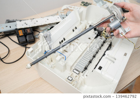 Handheld printer roller and motor above an open, disassembled printer on a desk, depicting repair, maintenance and parts replacement by a technician in an office or workshop setting. Handheld printer roller and motor above an open, disassembled printer on a desk, depicting repair, maintenance and parts replacement by a technician in an office or workshop setting. 139462792