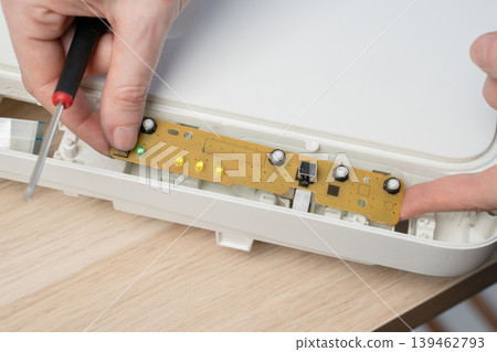 Close-up of a hand using a screwdriver to remove a screw from an exposed circuit board inside a printer device, illustrating electronic repair, DIY maintenance and small electronics troubleshooting. Close-up of a hand using a screwdriver to remove a screw from an exposed circuit board inside a printer device, illustrating electronic repair, DIY maintenance and small electronics troubleshooting. 139462793