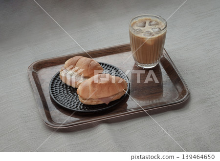 A wooden tray holds a refreshing iced latte in a textured glass, paired with two spinach and ham truffle salad rolls served on a decorative black plate. A wooden tray holds a refreshing iced latte in a textured glass, paired with two spinach and ham truffle salad rolls served on a decorative black plate. 139464650