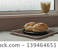 Two ham and truffle salad sandwiches sit on a dark wooden tray, accompanied by a ribbed glass of iced latte perched on the windowsill. The rich coffee colors offer a warm contrast to the soft, grey 139464651
