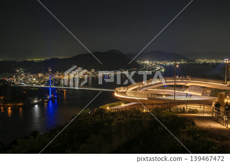 A view of the Kanmon Bridge and the nightscape of Shimonoseki from the observation deck at Hinoyama Park. The illuminated bridge and the lights of the city are dazzling. A view of the Kanmon Bridge and the nightscape of Shimonoseki from the observation deck at Hinoyama Park. The illuminated bridge and the lights of the city are dazzling. 139467472