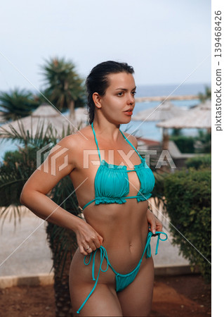 Pretty woman posing confidently in turquoise bikini with glowing bronzed healthy summer appearance. Pretty model showing smooth radiant body texture during tropical resort vacation beside palm Pretty woman posing confidently in turquoise bikini with glowing bronzed healthy summer appearance. Pretty model showing smooth radiant body texture during tropical resort vacation beside palm 139468426