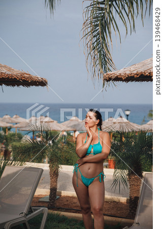 Female model standing beside tropical resort with glowing healthy bronzed summer complexion outdoors. Beautiful female showing smooth radiant body texture during luxury vacation near seaside palms and Female model standing beside tropical resort with glowing healthy bronzed summer complexion outdoors. Beautiful female showing smooth radiant body texture during luxury vacation near seaside palms and 139468429