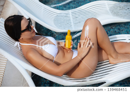 Attractive woman applying sunscreen lotion on smooth glowing legs beside swimming pool outdoors. Attractive female showing healthy radiant skin texture during luxury summer vacation near resort Attractive woman applying sunscreen lotion on smooth glowing legs beside swimming pool outdoors. Attractive female showing healthy radiant skin texture during luxury summer vacation near resort 139468451