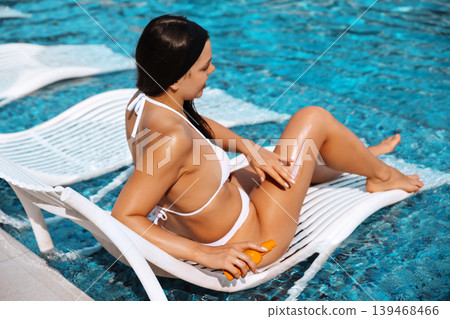 Beautiful woman holding sunscreen lotion with radiant bronzed skin beside luxury swimming pool. Beautiful female relaxing on floating lounge chair showing smooth glowing body texture during tropical Beautiful woman holding sunscreen lotion with radiant bronzed skin beside luxury swimming pool. Beautiful female relaxing on floating lounge chair showing smooth glowing body texture during tropical 139468466