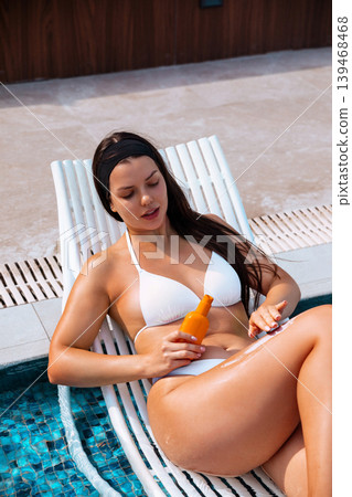 Pretty woman applying sunscreen lotion on smooth glowing legs beside swimming pool naturally. Pretty model holding skincare bottle while showing radiant healthy skin texture during luxury tropical Pretty woman applying sunscreen lotion on smooth glowing legs beside swimming pool naturally. Pretty model holding skincare bottle while showing radiant healthy skin texture during luxury tropical 139468468