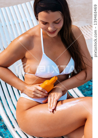 Attractive woman spreading sunscreen cream on radiant smooth legs beside luxury pool outdoors. Attractive female applying sun cream and holding skincare bottle while showing healthy glowing body Attractive woman spreading sunscreen cream on radiant smooth legs beside luxury pool outdoors. Attractive female applying sun cream and holding skincare bottle while showing healthy glowing body 139468469