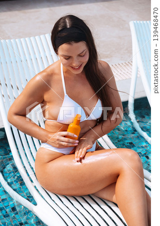 Young woman using sunscreen lotion with glowing healthy complexion beside tropical swimming pool outdoors. Beautiful female applying sun cream and holding skincare bottle while showing smooth radiant Young woman using sunscreen lotion with glowing healthy complexion beside tropical swimming pool outdoors. Beautiful female applying sun cream and holding skincare bottle while showing smooth radiant 139468470