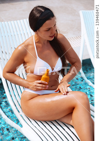 Female model applying sunscreen cream on smooth bronzed legs beside swimming pool naturally. Beautiful female holding skincare bottle while showing radiant healthy skin texture during tropical luxury Female model applying sunscreen cream on smooth bronzed legs beside swimming pool naturally. Beautiful female holding skincare bottle while showing radiant healthy skin texture during tropical luxury 139468471