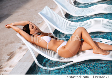 Beautiful woman relaxing on floating pool chair with glowing bronzed summer skin outdoors. Beautiful female posing in white bikini showing smooth radiant healthy body texture beside luxury swimming Beautiful woman relaxing on floating pool chair with glowing bronzed summer skin outdoors. Beautiful female posing in white bikini showing smooth radiant healthy body texture beside luxury swimming 139468475