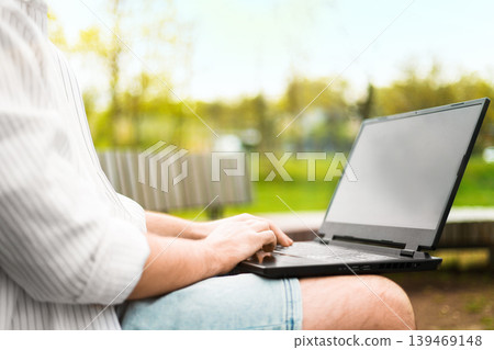 Man working on a laptop outdoors in a green park. Close-up of hands typing on a computer. Concept of remote work, freelancing, and digital nomad lifestyle in spring. 139469148