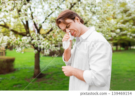 Man rubbing his itchy eyes with a tissue while standing in a park with blooming trees. Concept of severe seasonal allergy, pollen sensitivity, and hay fever during spring. 139469154