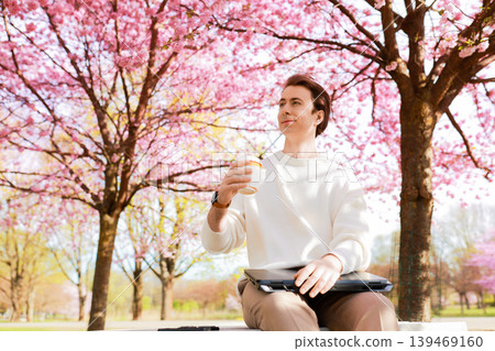 Smiling man sitting on a park bench with a laptop and coffee under blooming cherry blossoms. Remote work, freelancing, and spring lifestyle concept in a scenic nature setting. Smiling man sitting on a park bench with a laptop and coffee under blooming cherry blossoms. Remote work, freelancing, and spring lifestyle concept in a scenic nature setting. 139469160