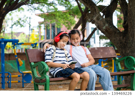 Two Asian student girls wearing headphones and using a smartphone together while sitting on a park bench, Back to school and digital lifestyle concept. Two Asian student girls wearing headphones and using a smartphone together while sitting on a park bench, Back to school and digital lifestyle concept. 139469226