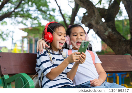 Two Asian student girls wearing headphones and using a smartphone together while sitting on a park bench, Back to school and digital lifestyle concept. Two Asian student girls wearing headphones and using a smartphone together while sitting on a park bench, Back to school and digital lifestyle concept. 139469227
