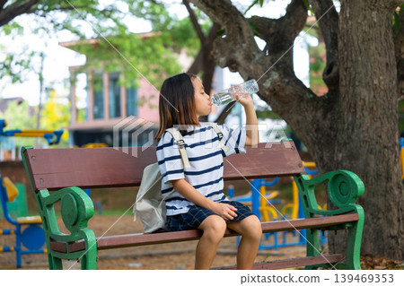 Cute Asian student girl sitting on a park bench and drinking water from a plastic bottle, Back to school and healthy lifestyle concept. Cute Asian student girl sitting on a park bench and drinking water from a plastic bottle, Back to school and healthy lifestyle concept. 139469353