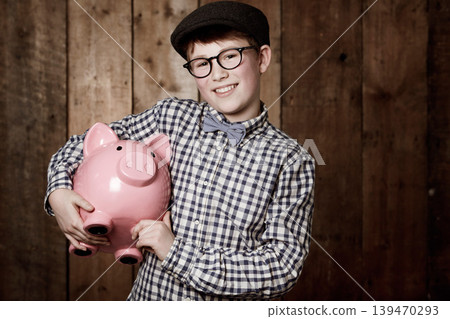 Young child, piggy bank or smile in portrait by wooden background, money saving or growth in youth in retro fashion. Boy, smile and face for coin banking and safe for bills with glasses by backdrop 139470293