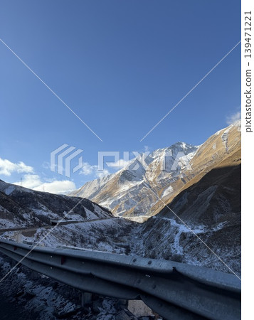 Mountain landscape with snowy peaks and clear blue sky, featuring textured guardrail in foreground Mountain landscape with snowy peaks and clear blue sky, featuring textured guardrail in foreground 139471221