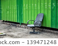Old black office chair abandoned on street near green metal wall. Concept of unemployment, discarded furniture and office closure. Urban decay and lonely object in city environment. 139471248