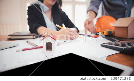 House model on table with group of two coworkers working with blueprint in office. co-working teamwork concept. House model on table with group of two coworkers working with blueprint in office. co-working teamwork concept. 139471288