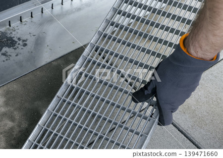 Close up of a hand in protective gloves removing a metal gutter grate, demonstrating simple DIY drainage maintenance. Close up of a hand in protective gloves removing a metal gutter grate, demonstrating simple DIY drainage maintenance. 139471660